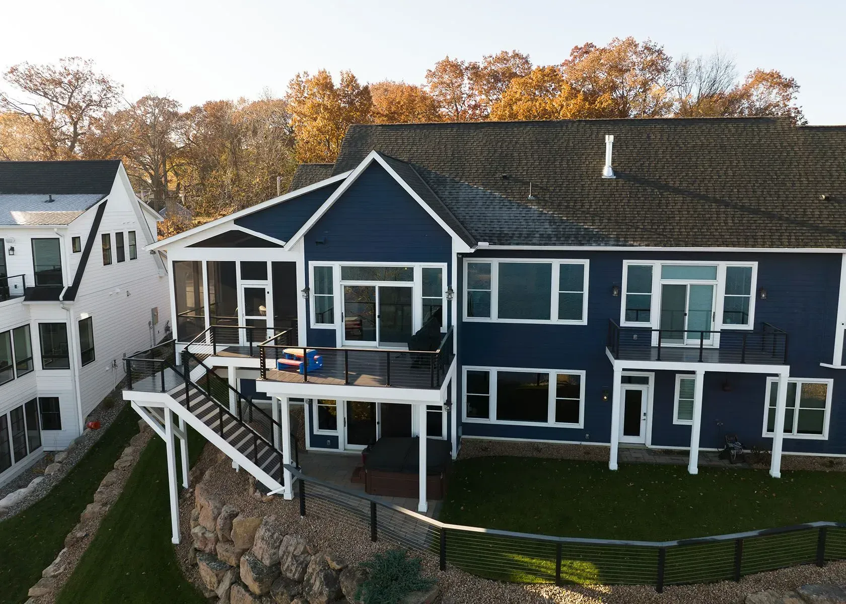 Blue and white multi-story houses with decks, stairs, and lake view, in a setting of green grass and fall foliage.