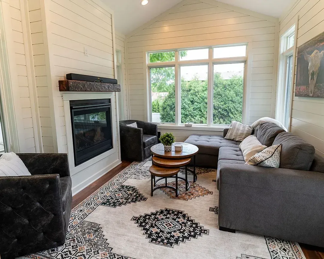 Cozy sunroom with fireplace, sectional sofa, and a patterned rug. White shiplap walls, and large windows with greenery outside.