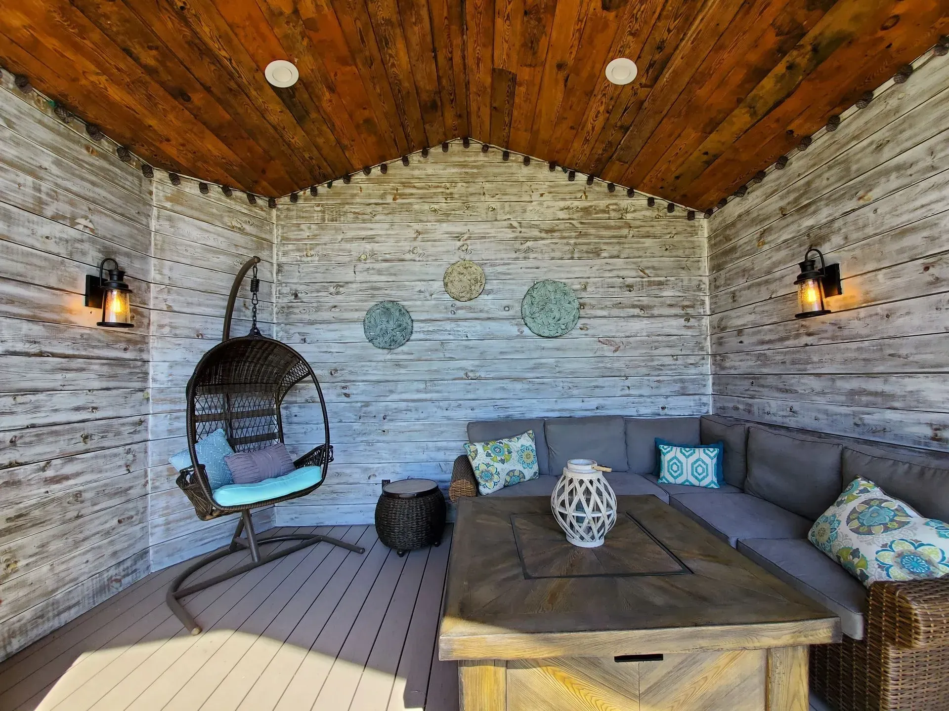 Covered outdoor patio with wood-paneled walls, gray sectional sofa, hanging chair, and wooden table.