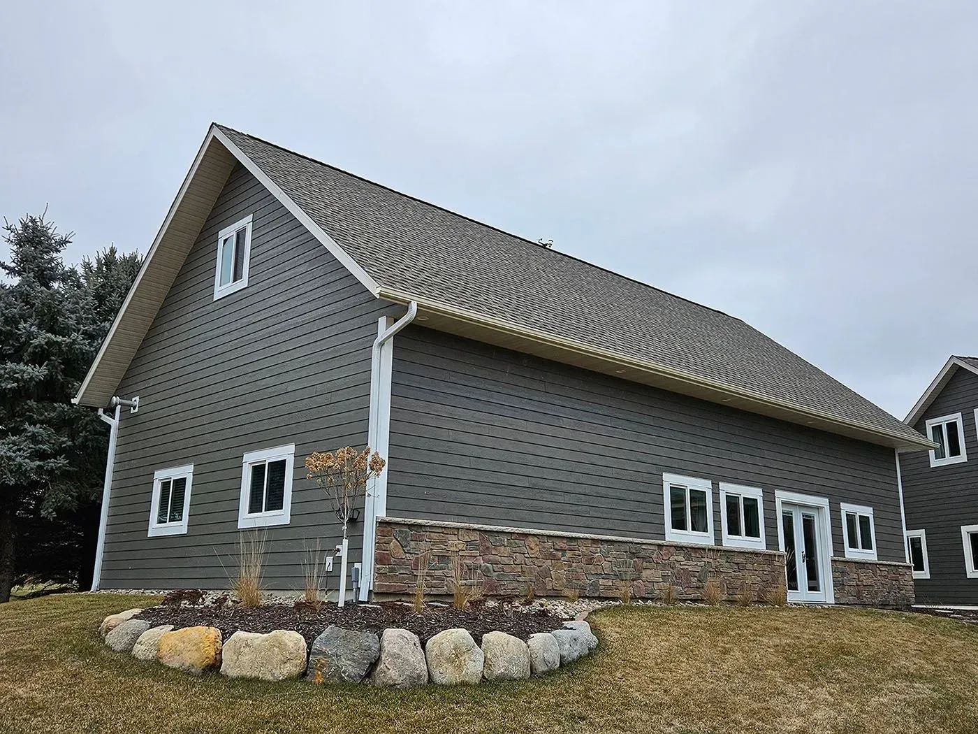 Gray building with stone base, white window frames, and brown roof under overcast sky.
