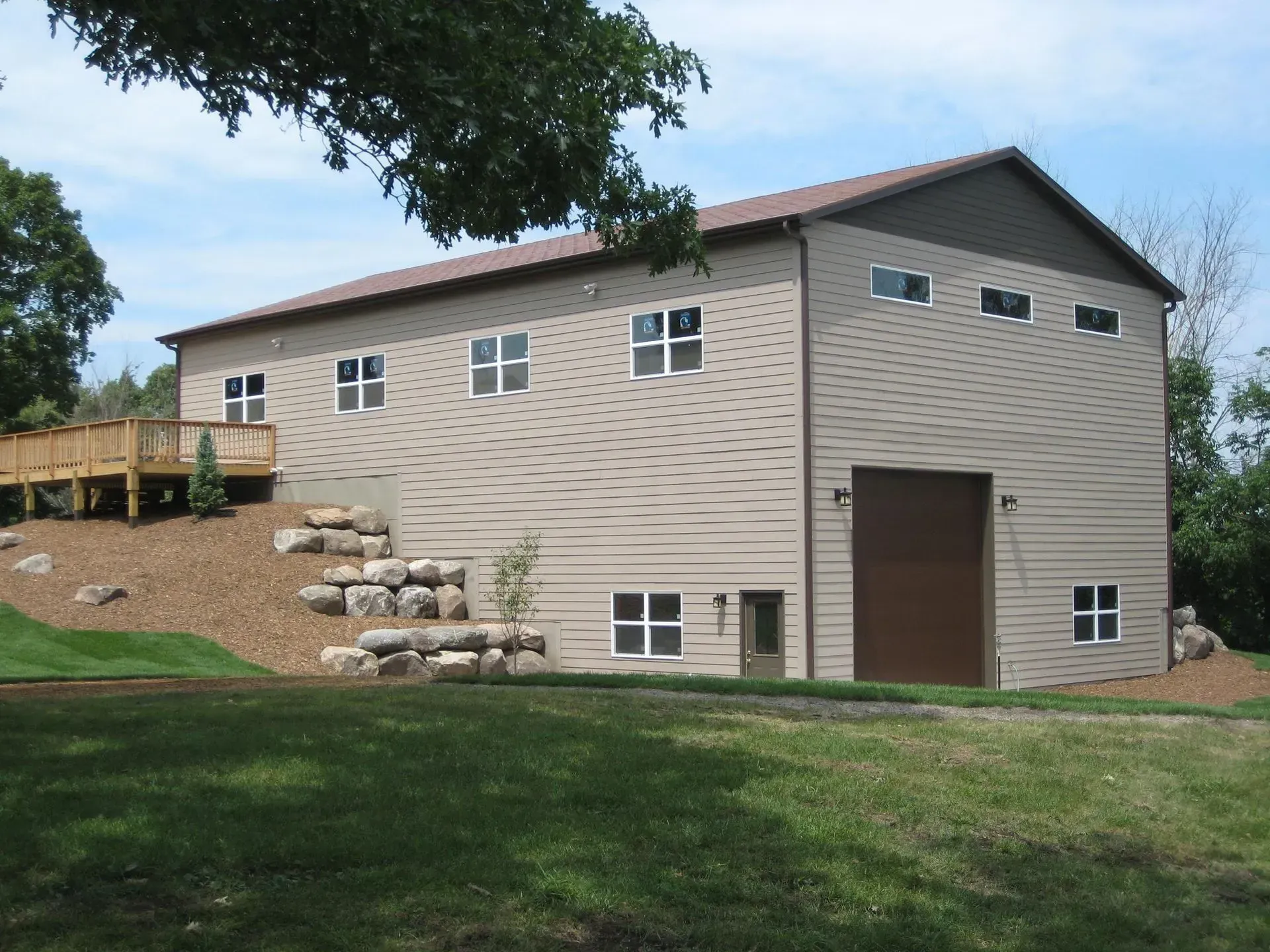 Two-story beige building with brown door, windows, and roof on a grassy hill; a wooden deck is visible.