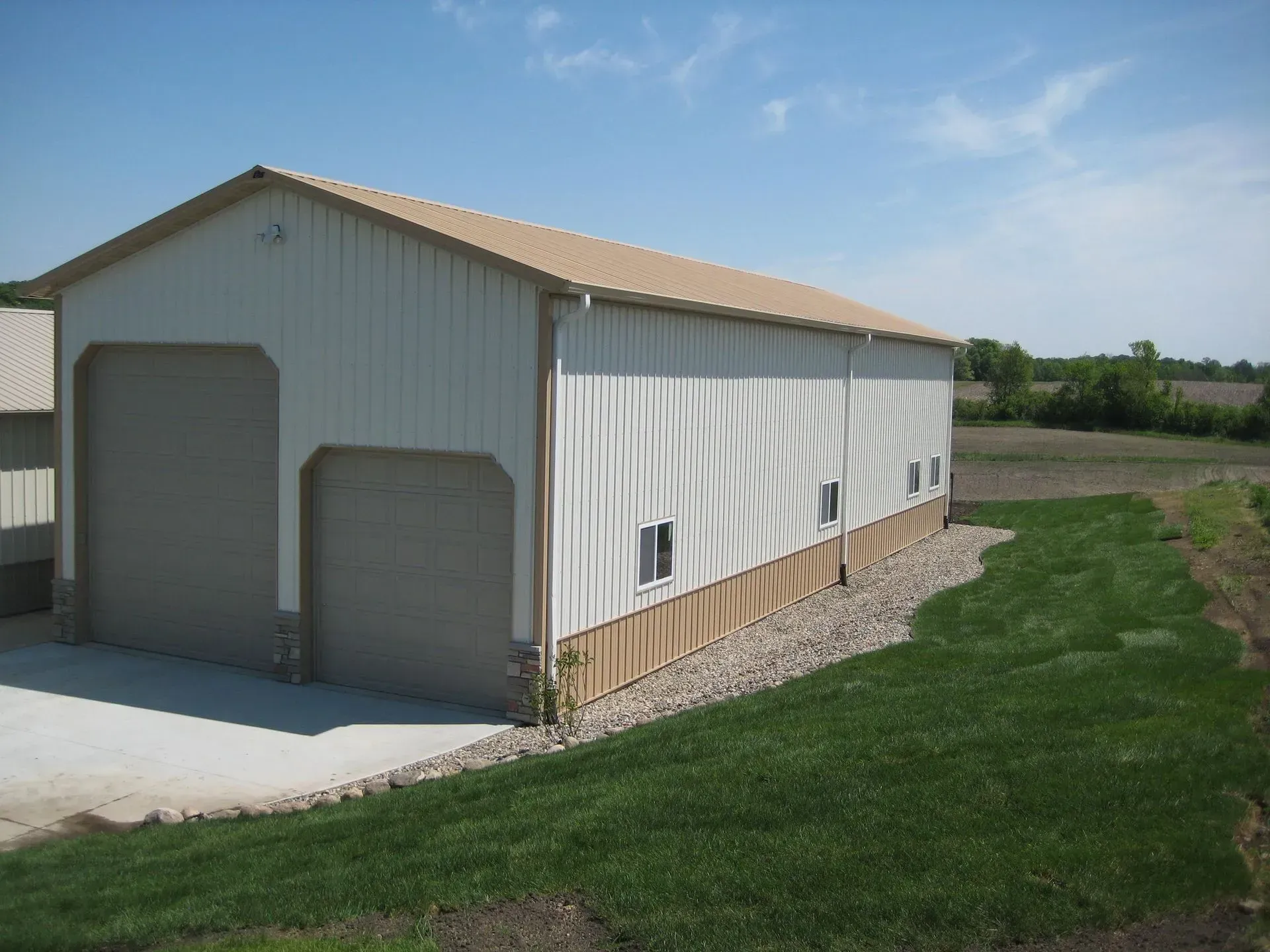 Tan and white metal building with two garage doors, brown trim, and a gravel base, on a grassy lot.