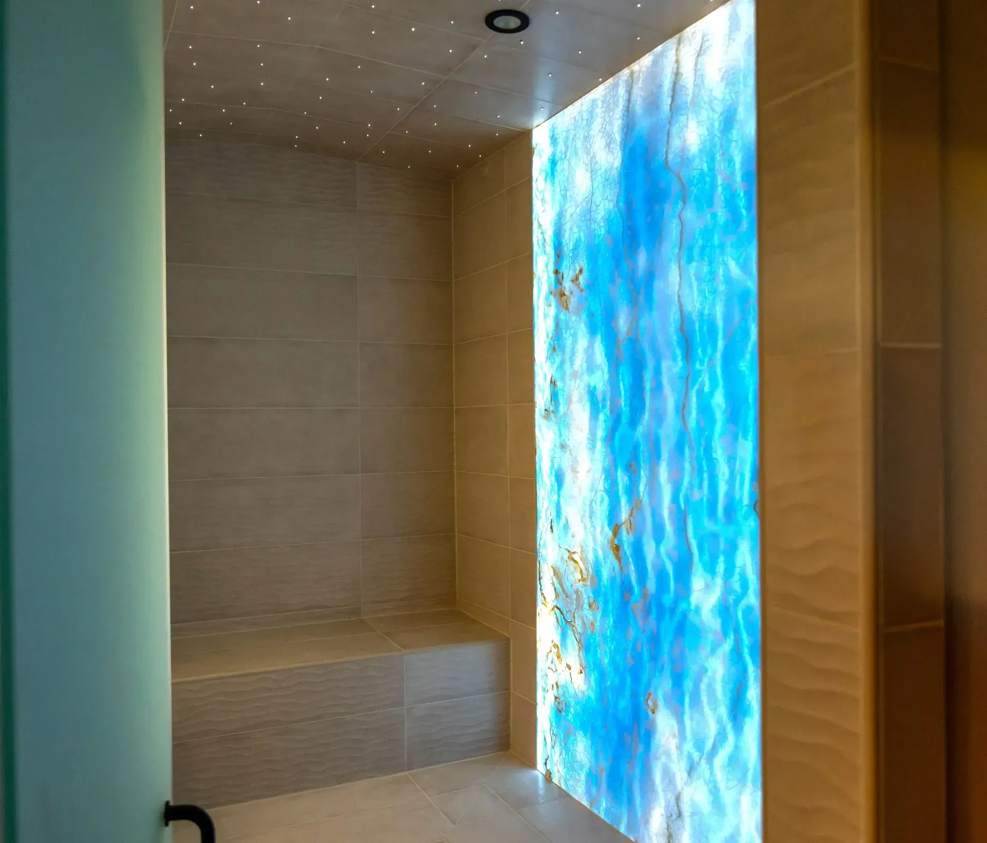 Shower stall with a blue illuminated wall, tan tiled walls, and a bench.