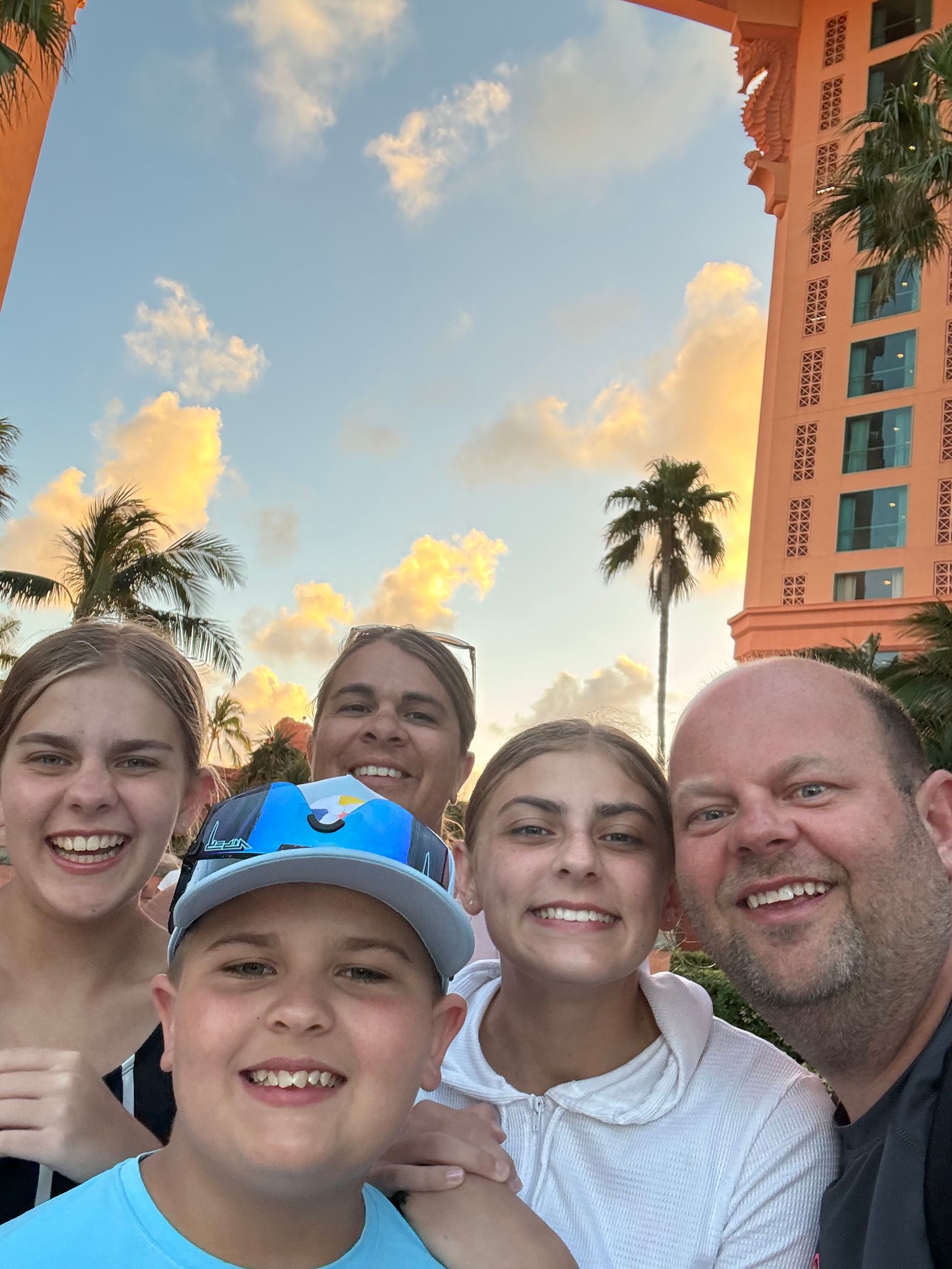 Family selfie at a sunny resort with palm trees and a tall orange building in the background