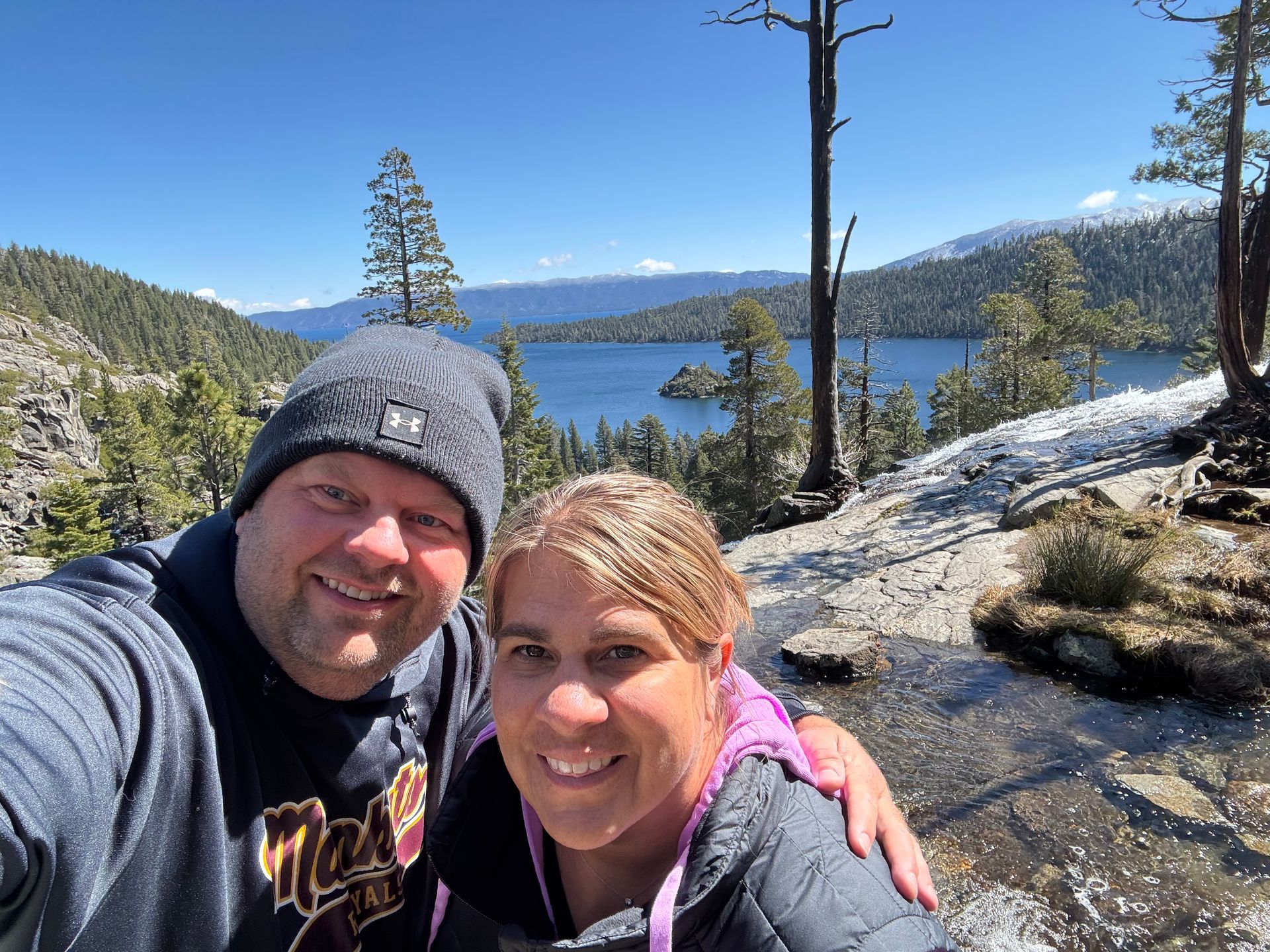 Smiling couple taking a selfie beside a mountain lake with snowy peaks and pine trees.