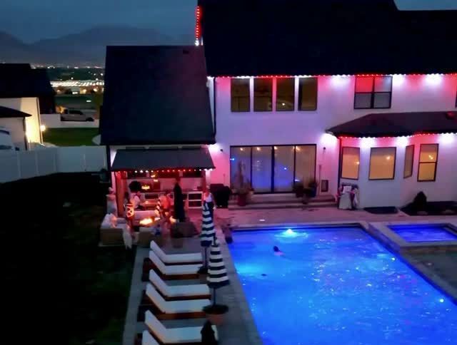 Nighttime poolside patio with blue-lit pool, striped loungers, and a house decorated with red lights.