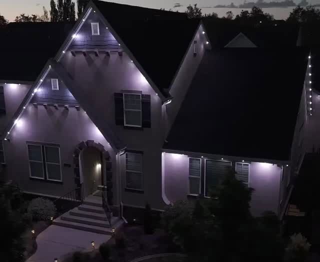 House at night with purple accent lights, lit windows, and string lights along the roofline.