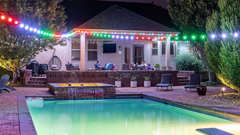 Backyard pool at dusk with colorful string lights and a house patio in the background