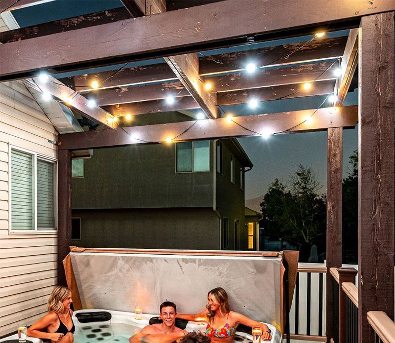 People relaxing in a lit backyard hot tub under a covered patio at dusk