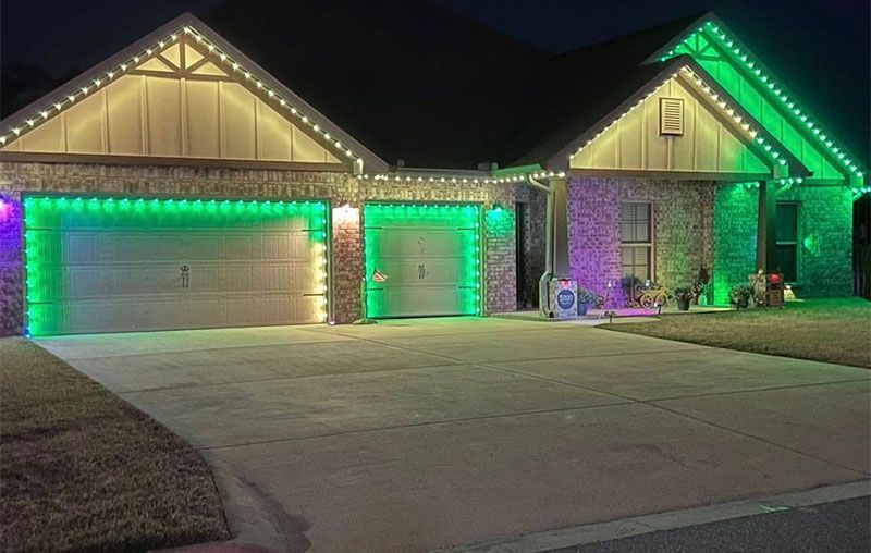Suburban house at night outlined in green and purple holiday lights with two garage doors and driveway