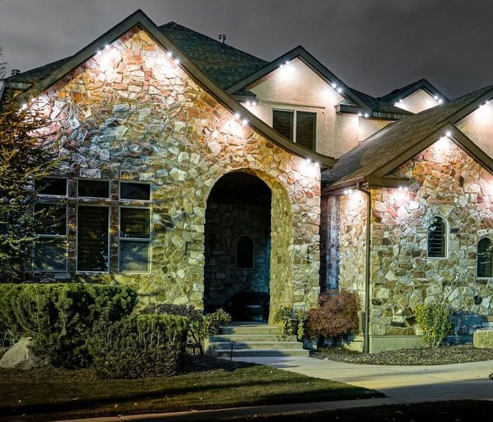 Stone house exterior at night, lit by warm porch lights and framed by shrubs.