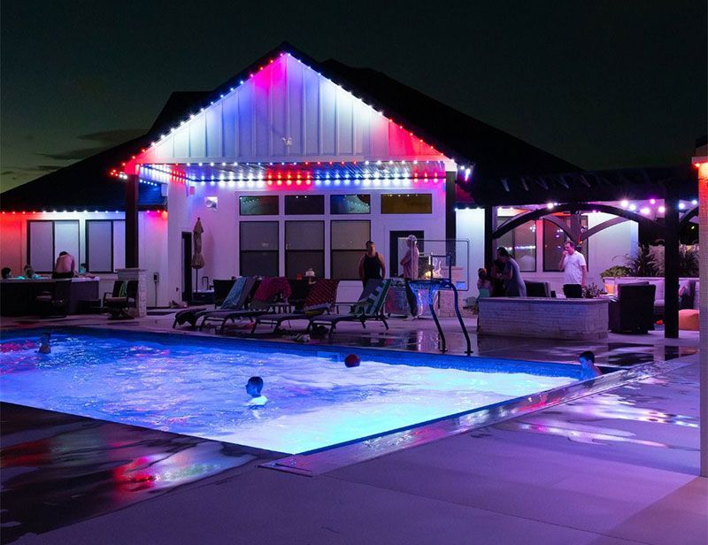 Nighttime backyard pool with colorful string lights and a lit house patio.