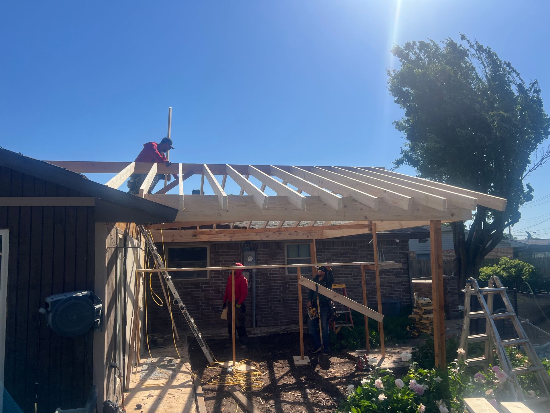 Construction workers building a wooden roof on a house extension under a bright blue sky.