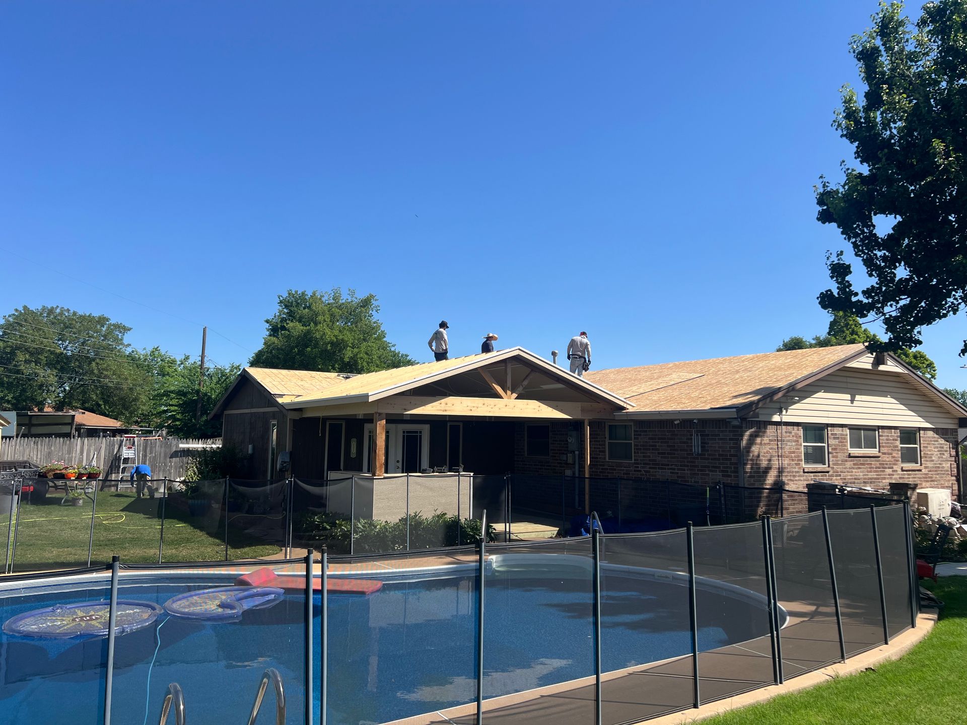 House under construction with workers on the roof; pool and yard in foreground.