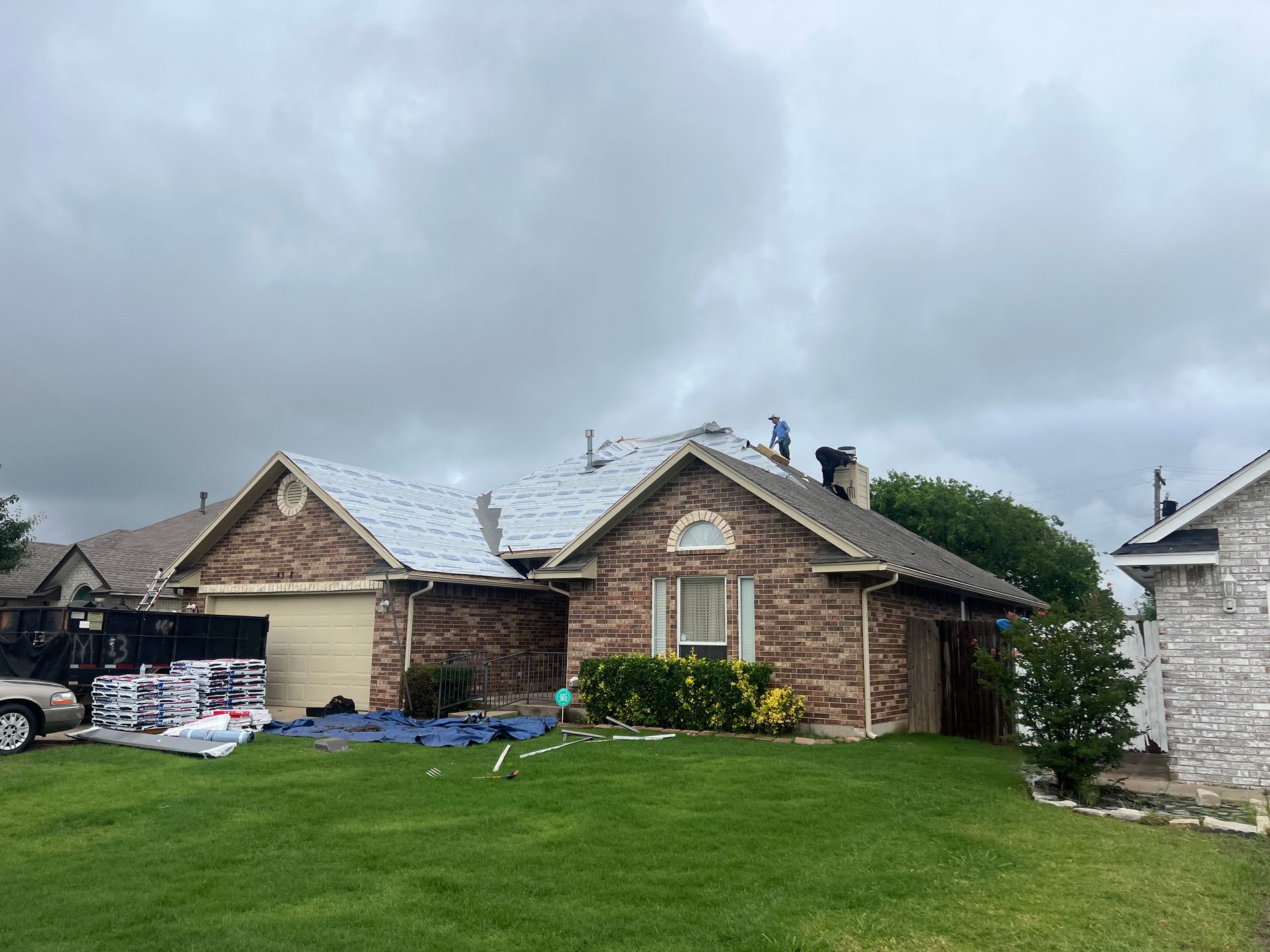 Roofers replacing shingles on a brick house under a cloudy sky.