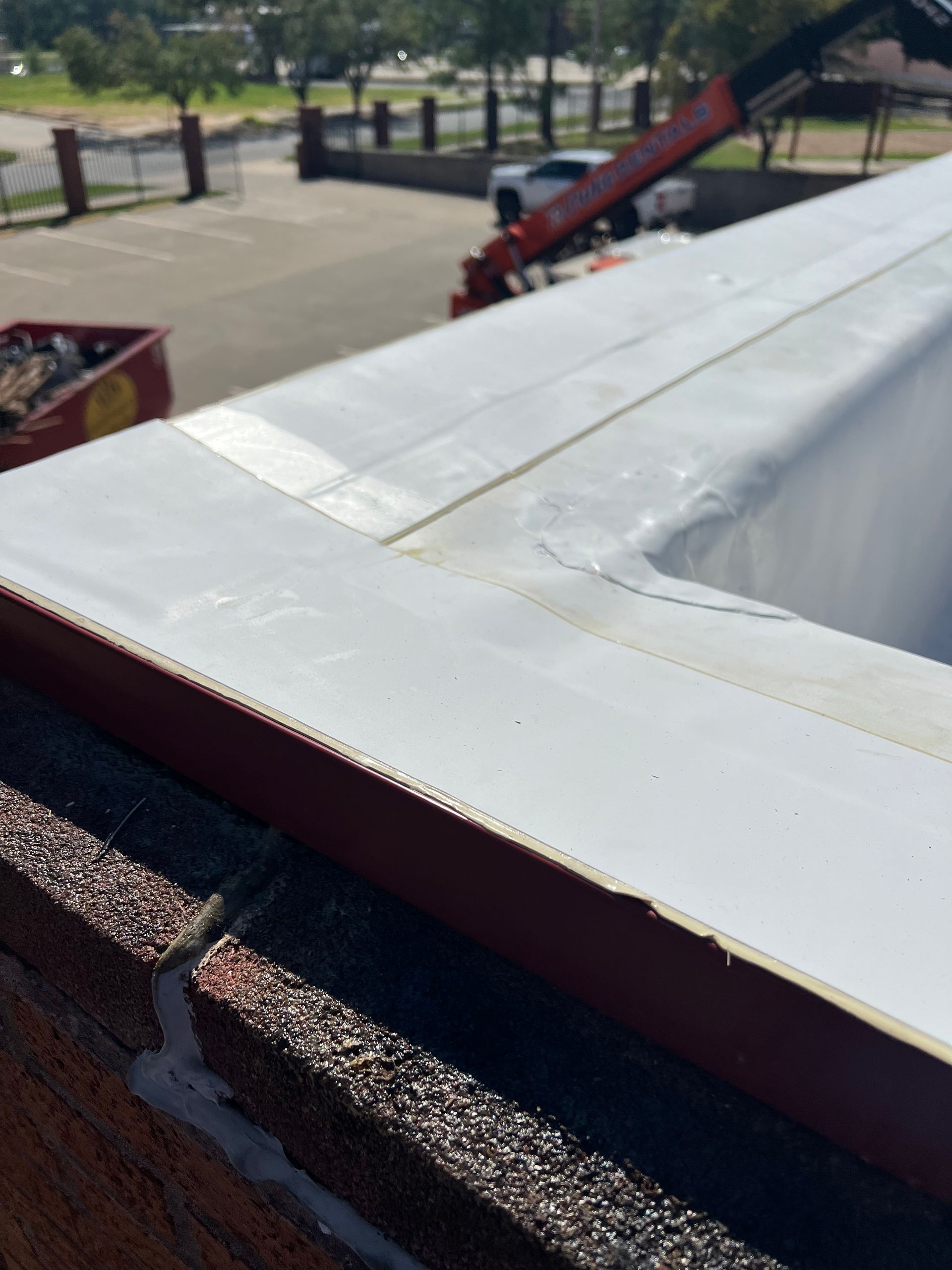 Close-up of white rooftop edge on brick building. A red trim and a dumpster are visible.