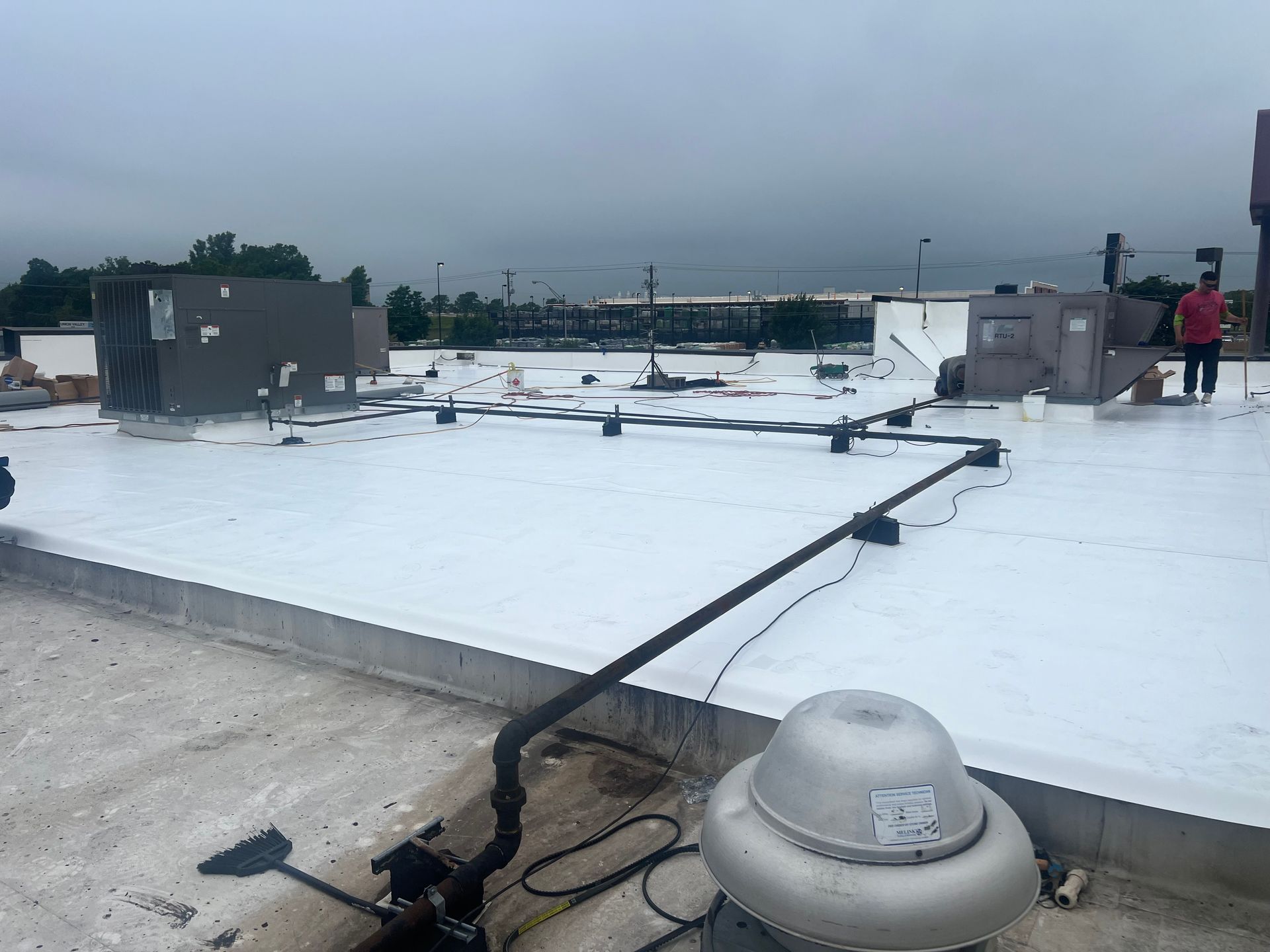 Rooftop with a white surface, various equipment, and a person in a red shirt standing near the edge under an overcast sky.