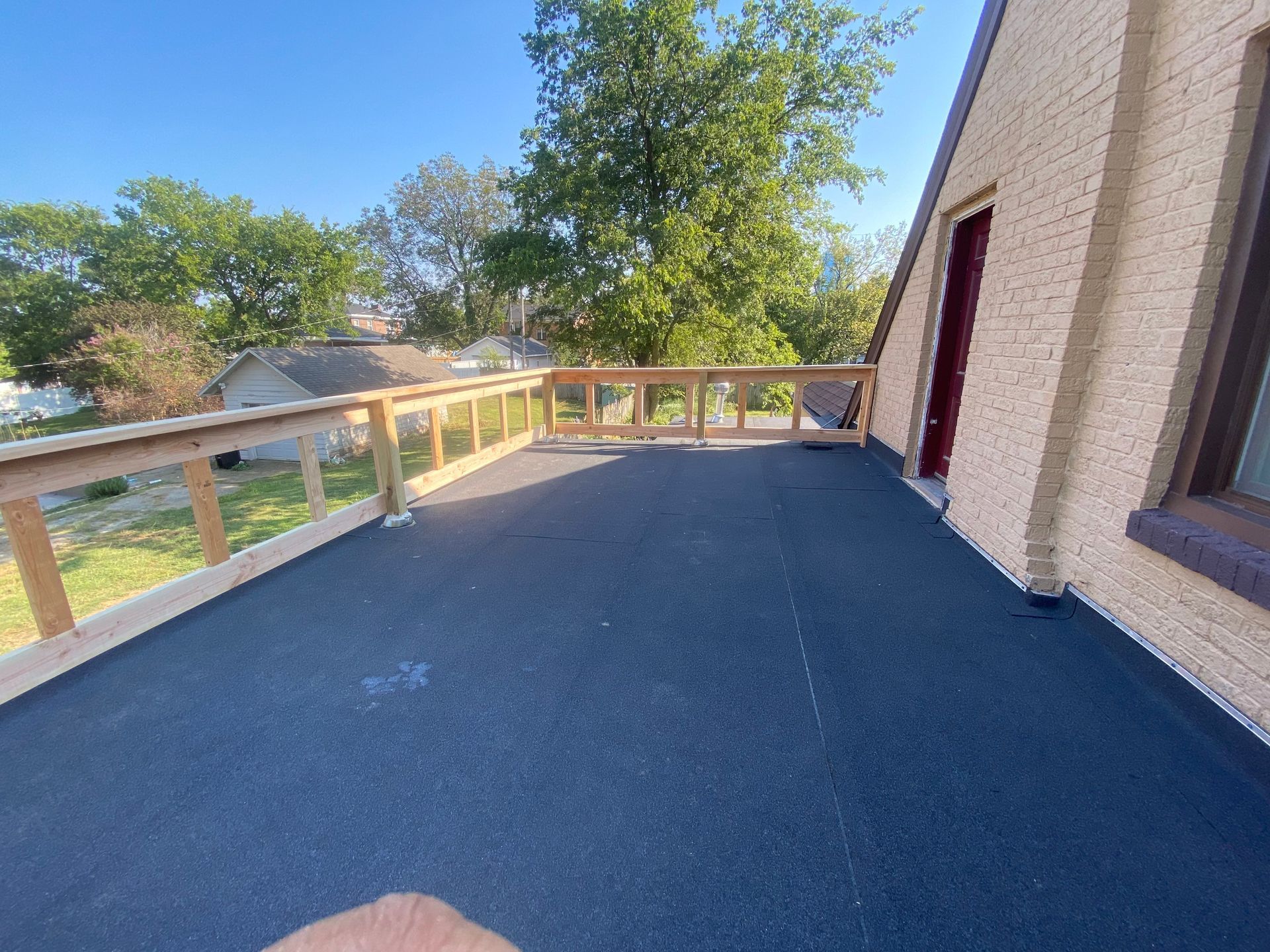 Exterior deck with black surface, wooden railings, and house, trees in background.