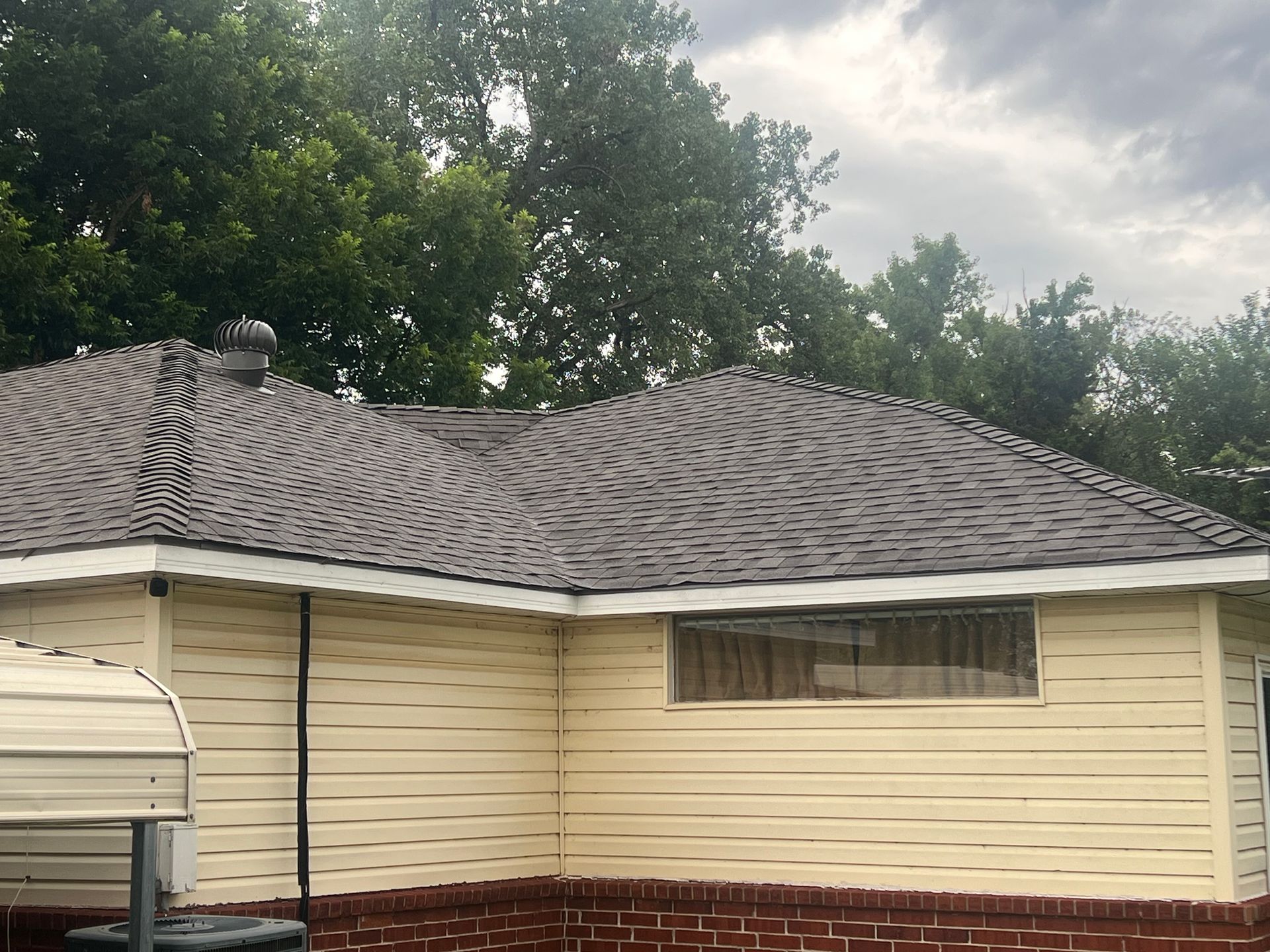Yellow-sided house with brown roof and brick base. Overcast sky, trees in the background.