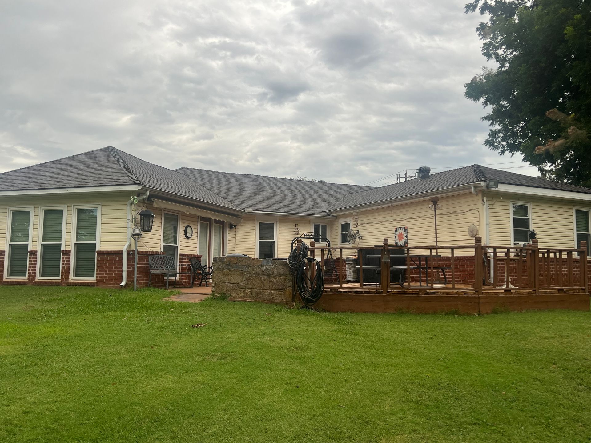 Back view of a one-story house with a wooden deck. Beige siding, red brick, and a dark shingled roof under a cloudy sky.