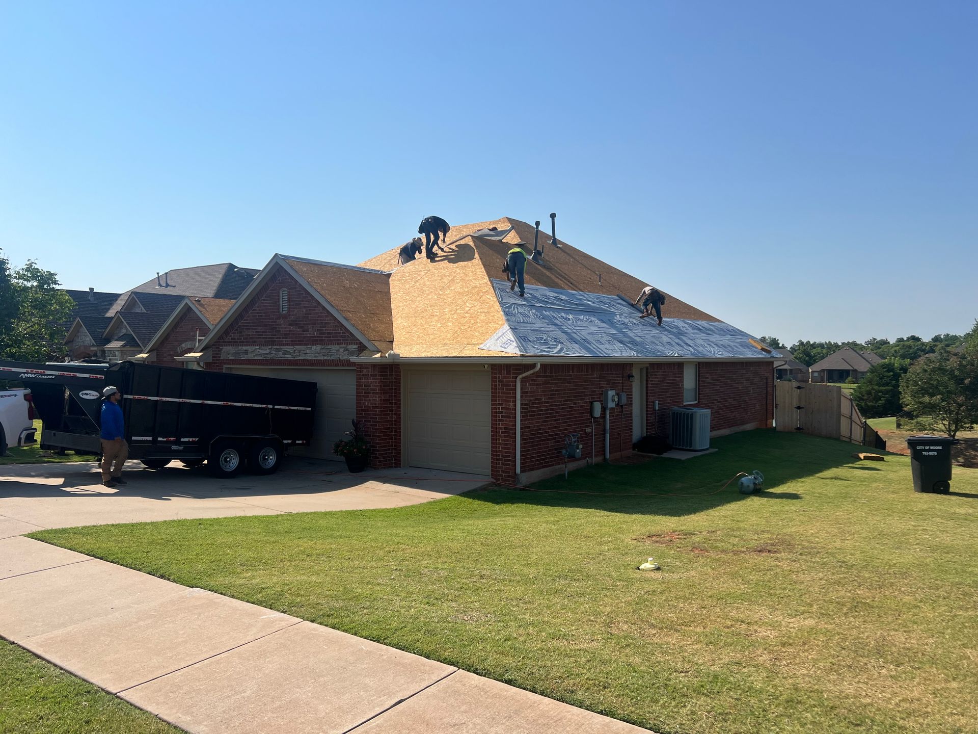 Roofers on a residential roof installing shingles on a sunny day. A truck with debris is parked nearby.