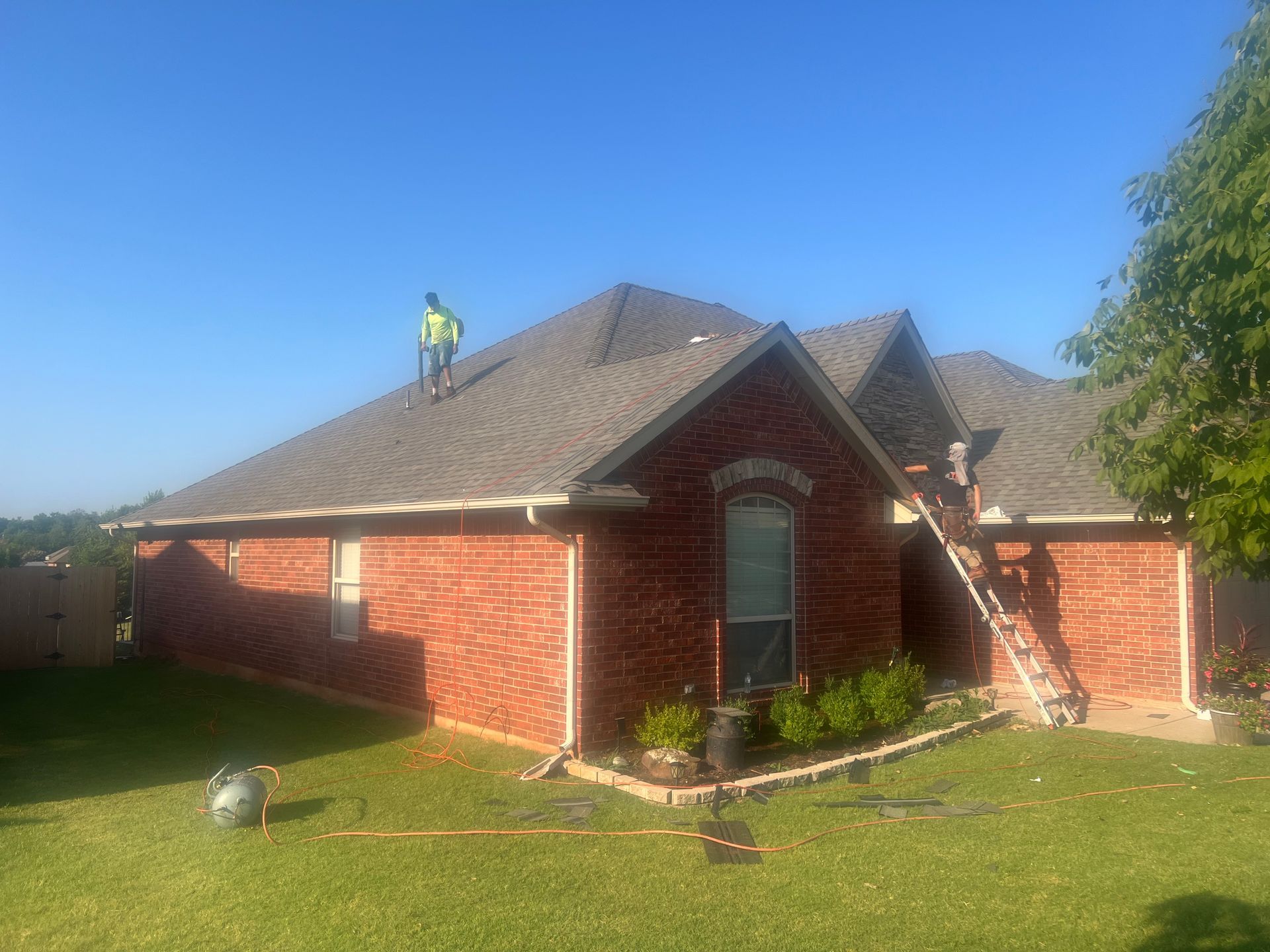 A worker on a roof of a brick house with a ladder. Blue sky, green grass.