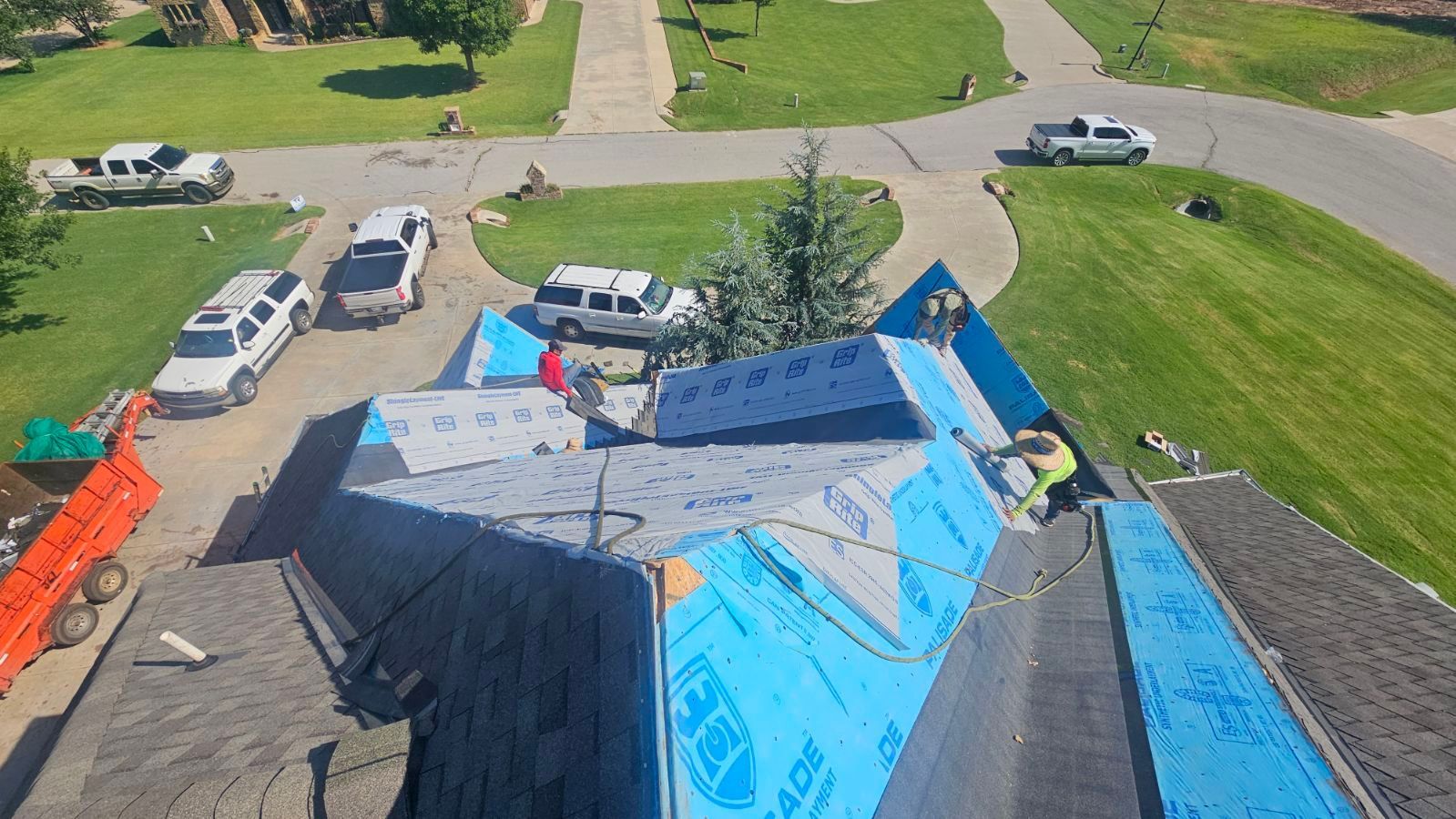 Roofers working on a house roof, blue underlayment visible, several trucks parked nearby.
