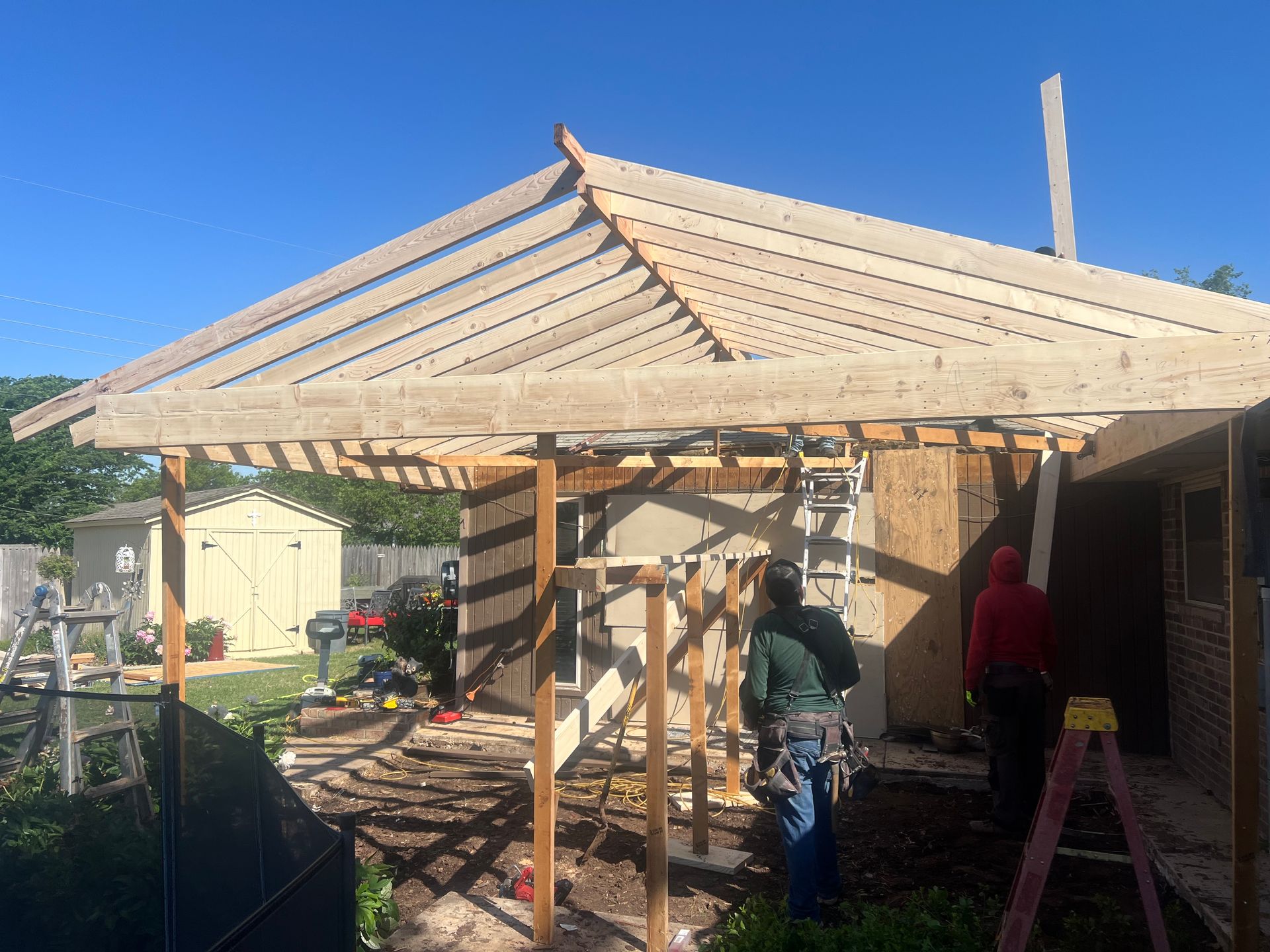 Construction of a wooden gazebo roof attached to a building, with workers, tools, and a sunny outdoor setting.