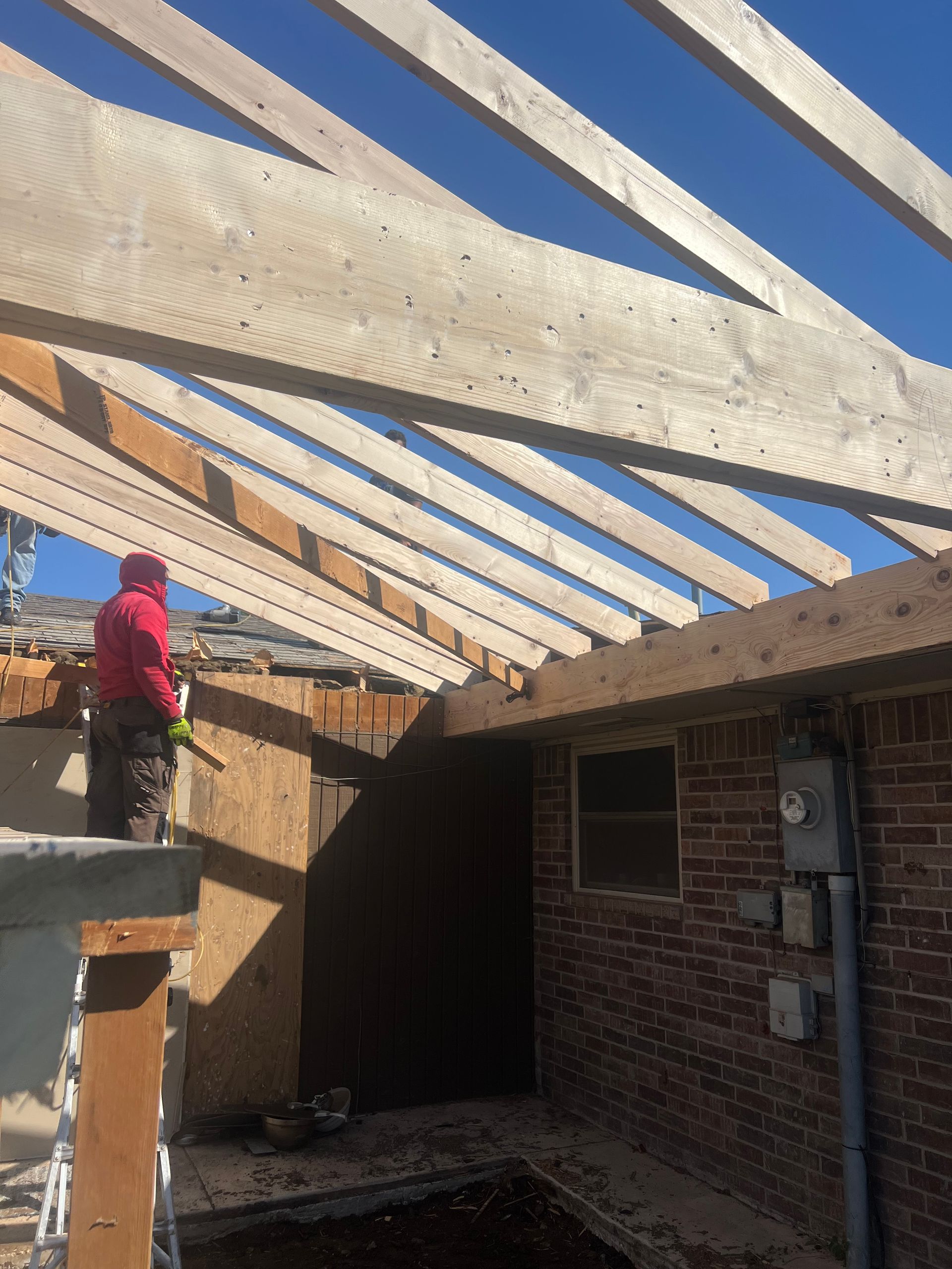 Construction worker on a partially built roof, with wood beams against a blue sky. Brick wall visible.