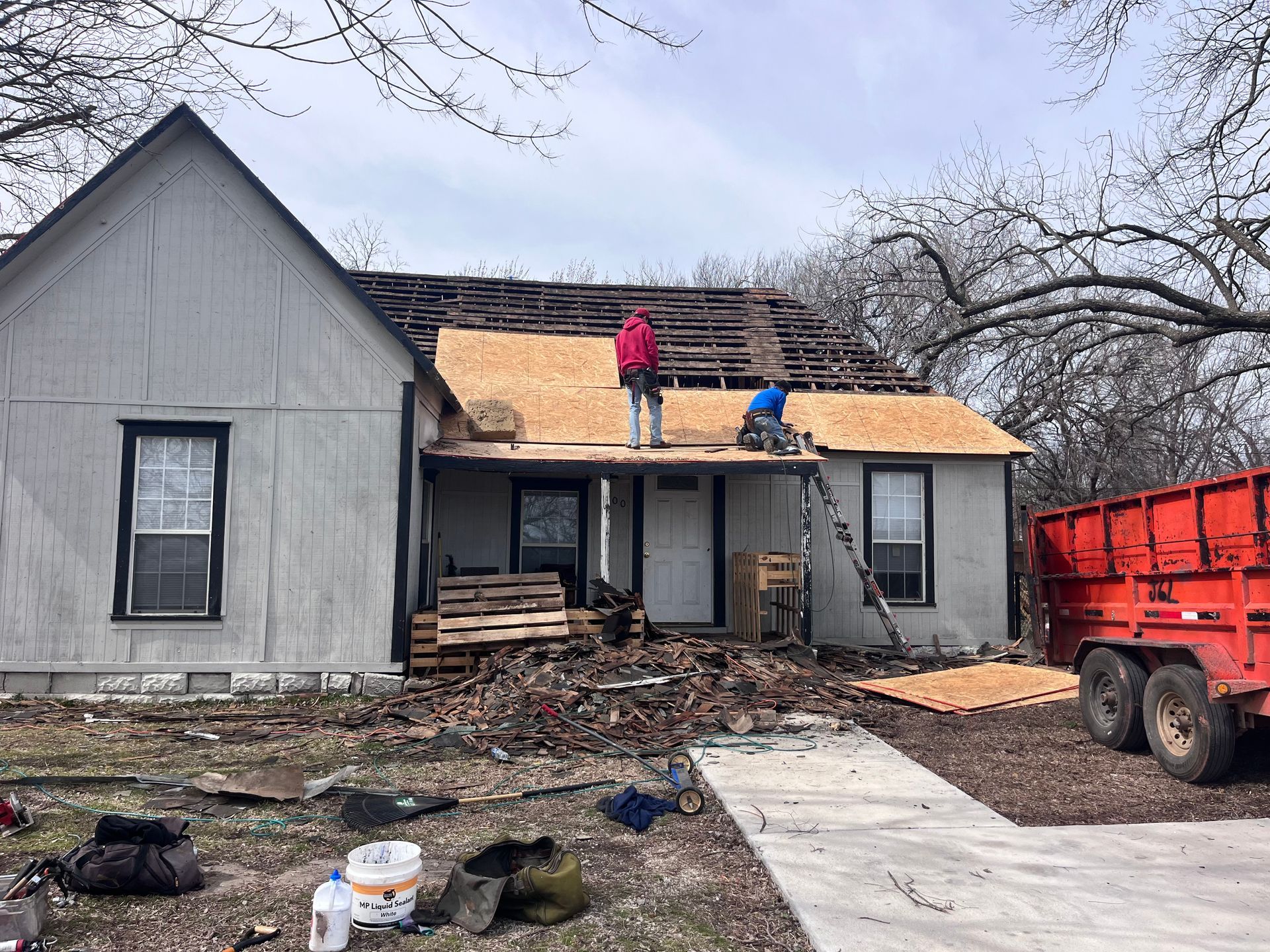 People on a house roof replacing shingles; debris on the ground; red trailer.