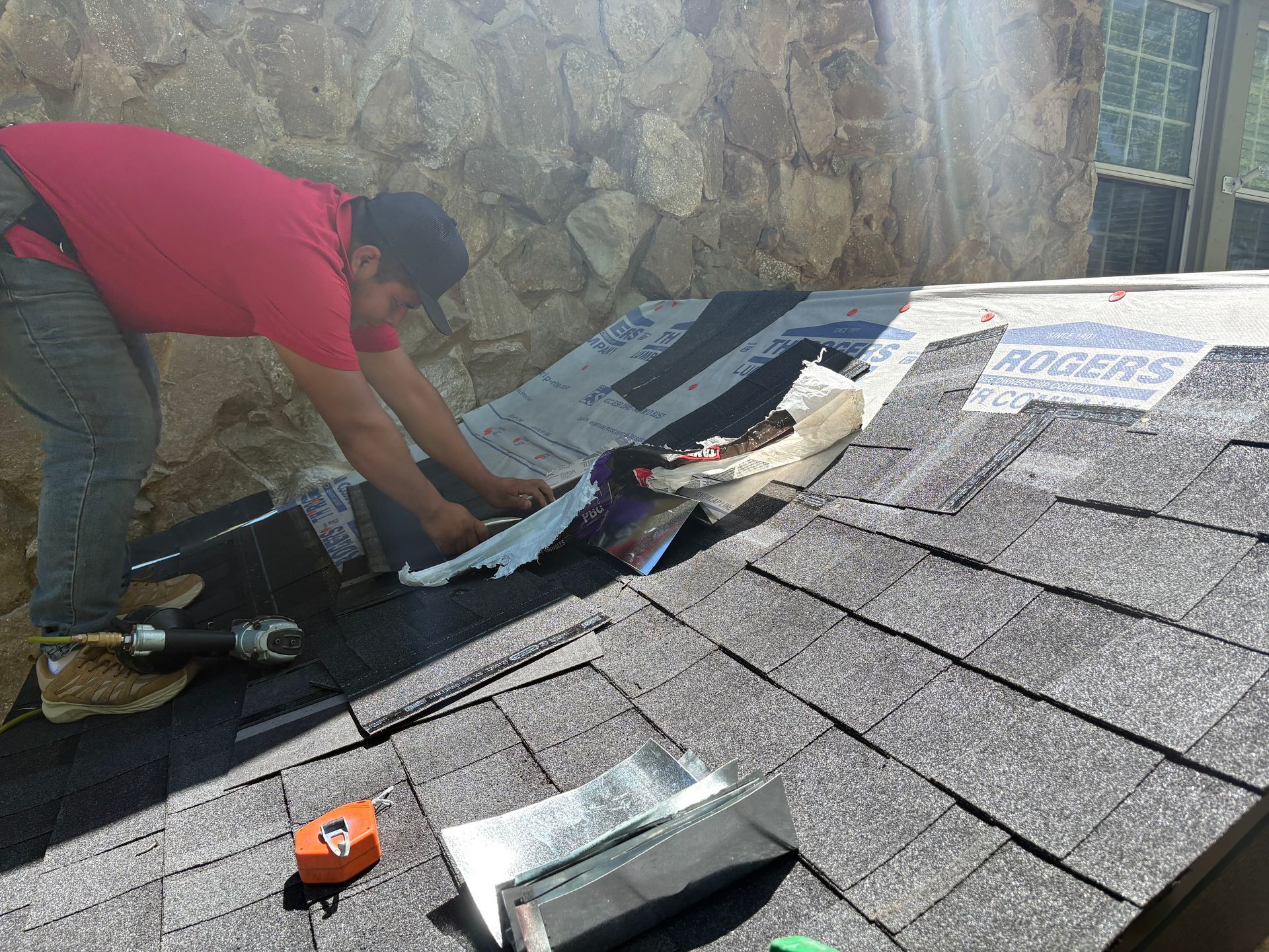 Roofer installing flashing on a shingled roof, next to a stone wall. He is wearing a red shirt and jeans.