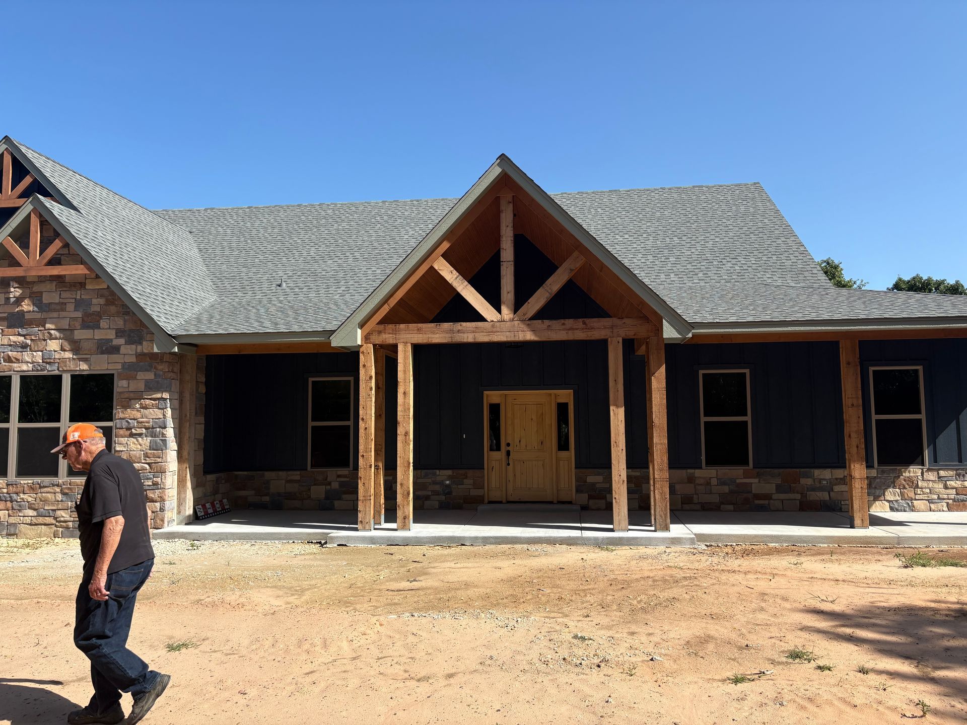 Man walks in front of a new home with stone accents, blue siding, and wooden entryway. Clear blue sky.