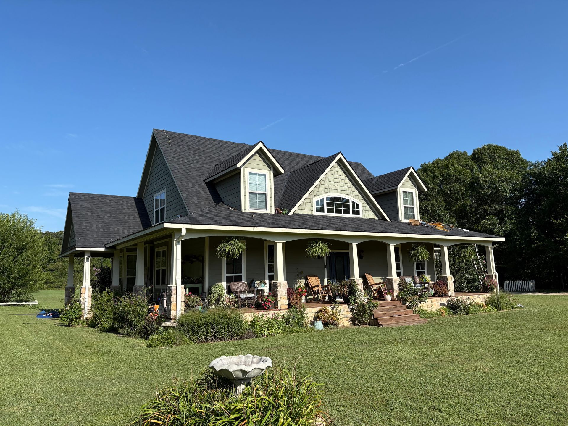 Farmhouse with wraparound porch, green siding, black roof, and manicured lawn under a clear blue sky.