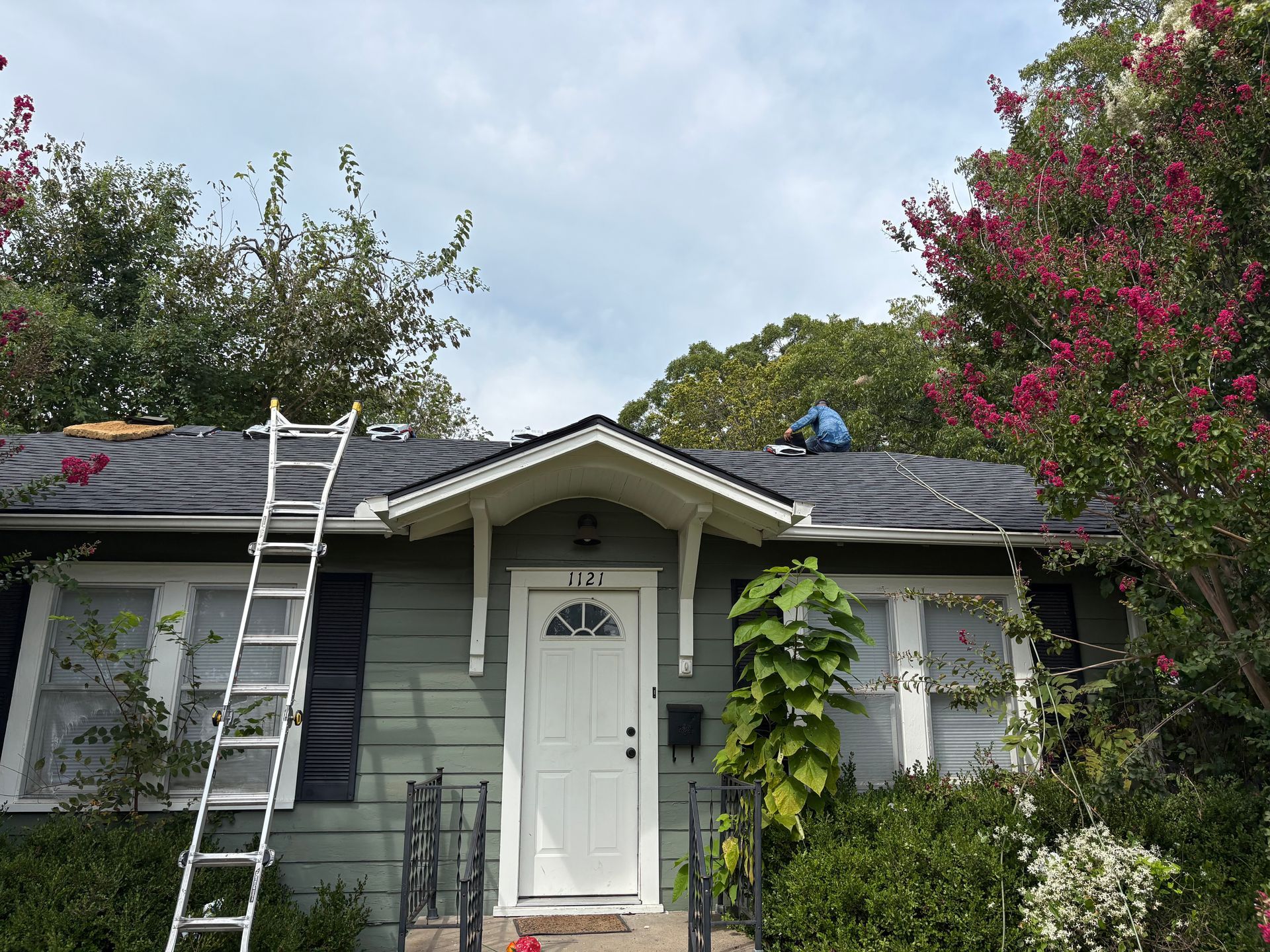 House with roof being worked on, ladder, foliage, cloudy sky.