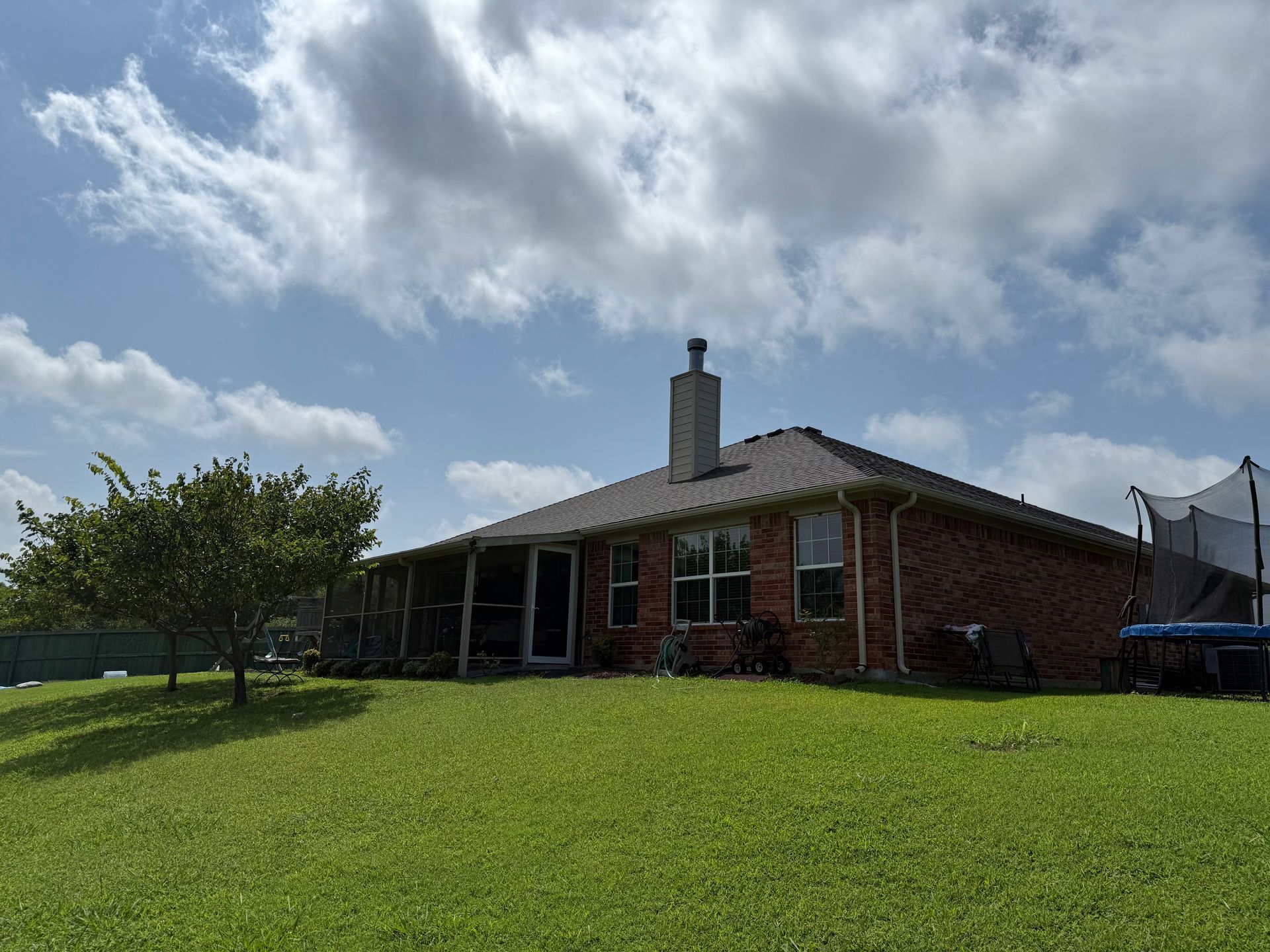 Backyard view of a brick house with a chimney, windows, a tree, and a trampoline under a cloudy sky.