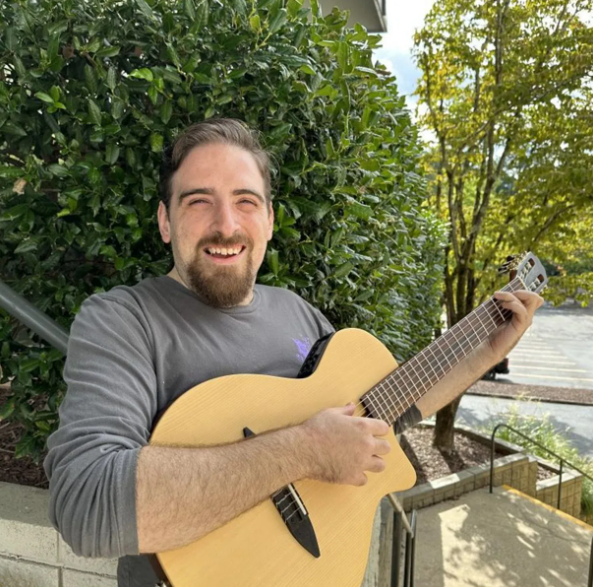 Man with a beard smiling, holding a light wood acoustic guitar outside, green foliage in the background.