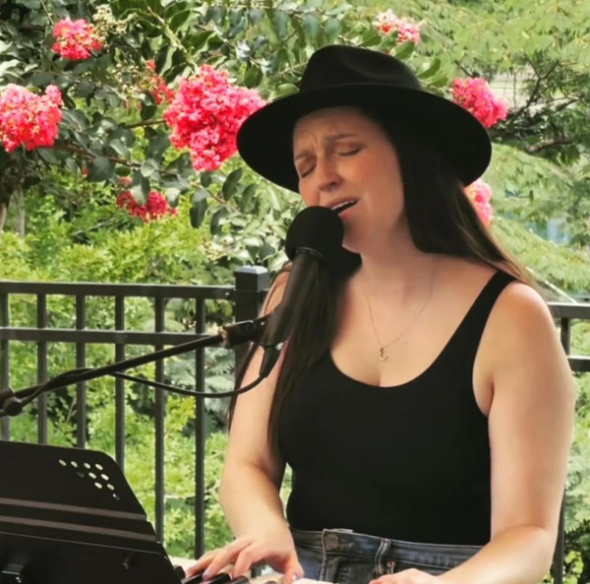 Woman in black hat singing into a microphone, playing a keyboard outdoors near pink flowers.