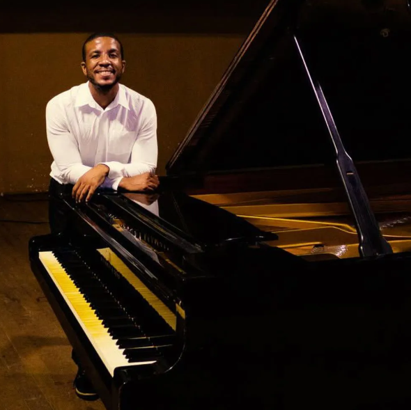 Man in white shirt leans on a grand piano, smiling. Interior shot with warm lighting.