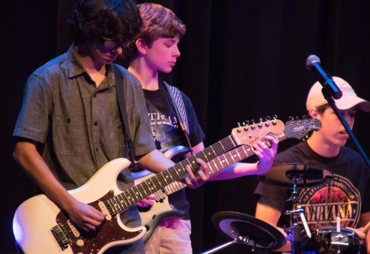 Three young musicians perform onstage. One plays drums, two play electric guitars. Dark background.