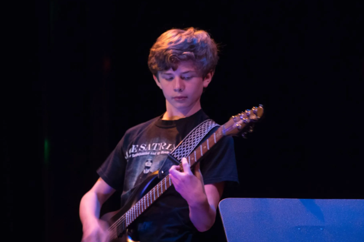 Teen playing electric guitar onstage, black background, bright lighting.
