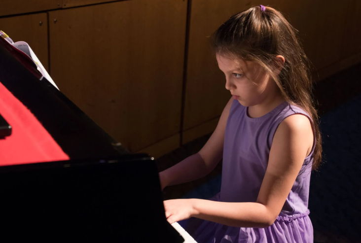 Girl in purple dress plays piano, focused expression, concert setting.