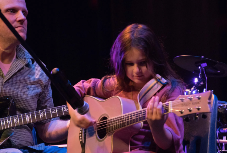 Young person playing an acoustic guitar onstage. Another person partially visible on the left.