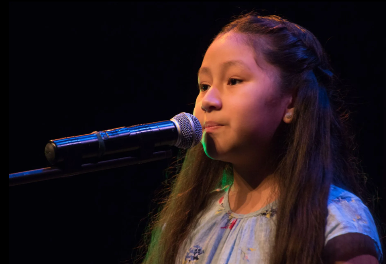 Young person singing into a microphone on stage, with long hair and a light-colored top.