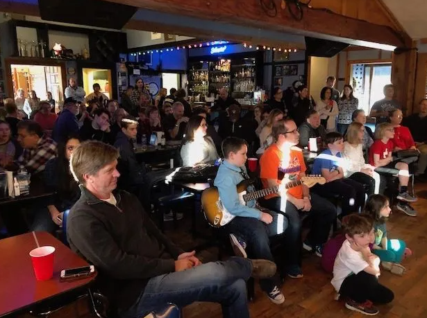 People at a bar, watching a performance. A boy sits with a guitar. Dark interior, some lights.