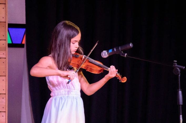 Young person playing a violin on stage, facing forward, with a microphone nearby.