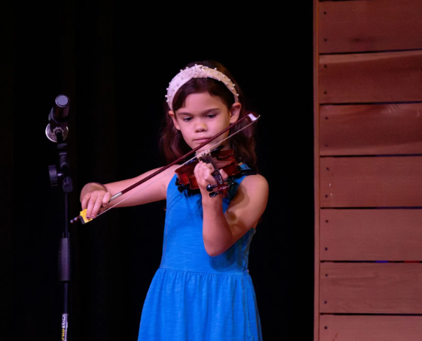 Young girl playing a violin on stage, wearing a blue dress and a headband.