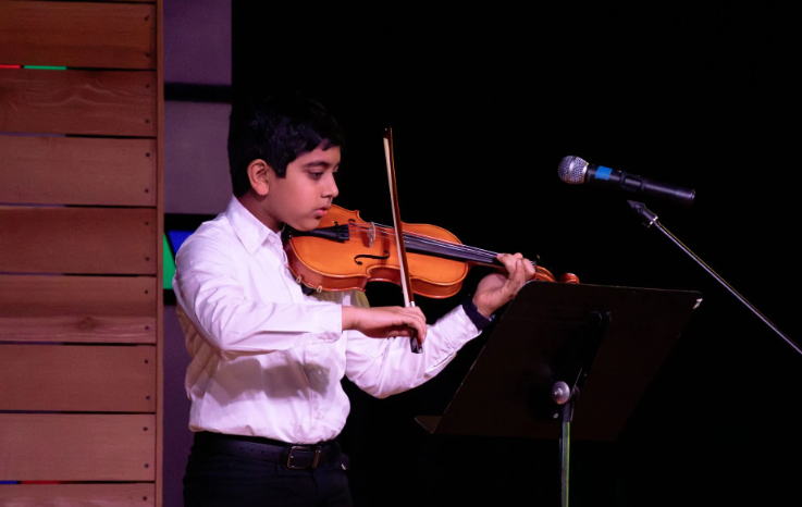 Boy in white shirt playing violin on stage with music stand.