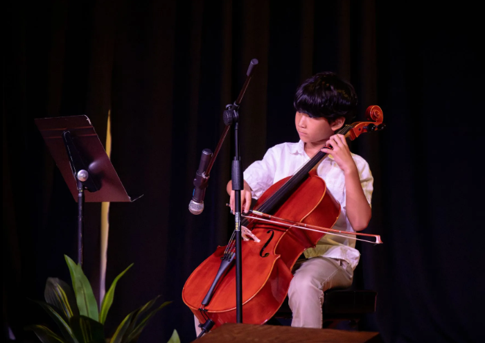 A young person playing a cello on stage under a spotlight, with a music stand and microphone nearby.