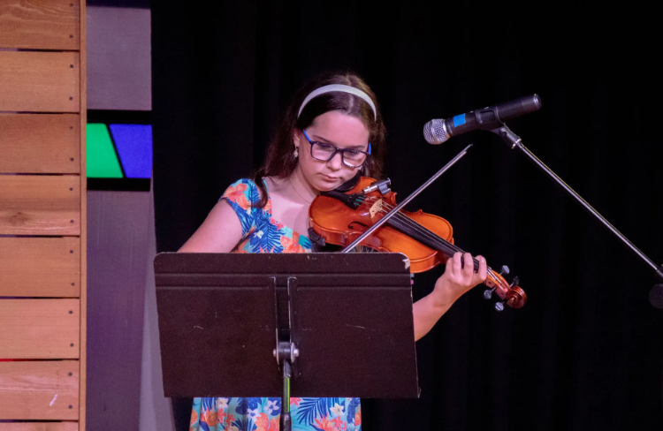 Young person playing a violin on stage, reading sheet music, with a microphone in front of them.