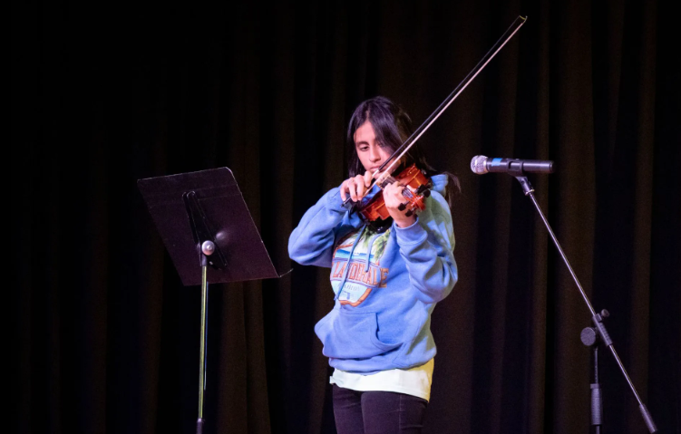 A person playing a violin on stage with a music stand and microphone. They wear a blue hoodie.